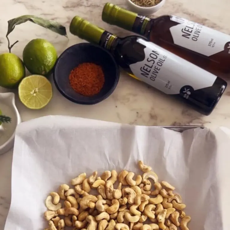 Ingredients for sweet and spicy cashews including raw cashews, maple syrup, rosemary, lime oil, and sea salt on white surface
