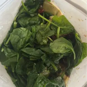 Reusable freezer bag with layered Tuscan chicken ingredients: cubed chicken, artichoke hearts, sun-dried tomatoes, spinach, herbs, and balsamic dressing