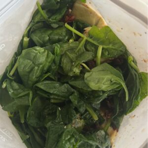 Reusable freezer bag with layered Tuscan chicken ingredients: cubed chicken, artichoke hearts, sun-dried tomatoes, spinach, herbs, and balsamic dressing